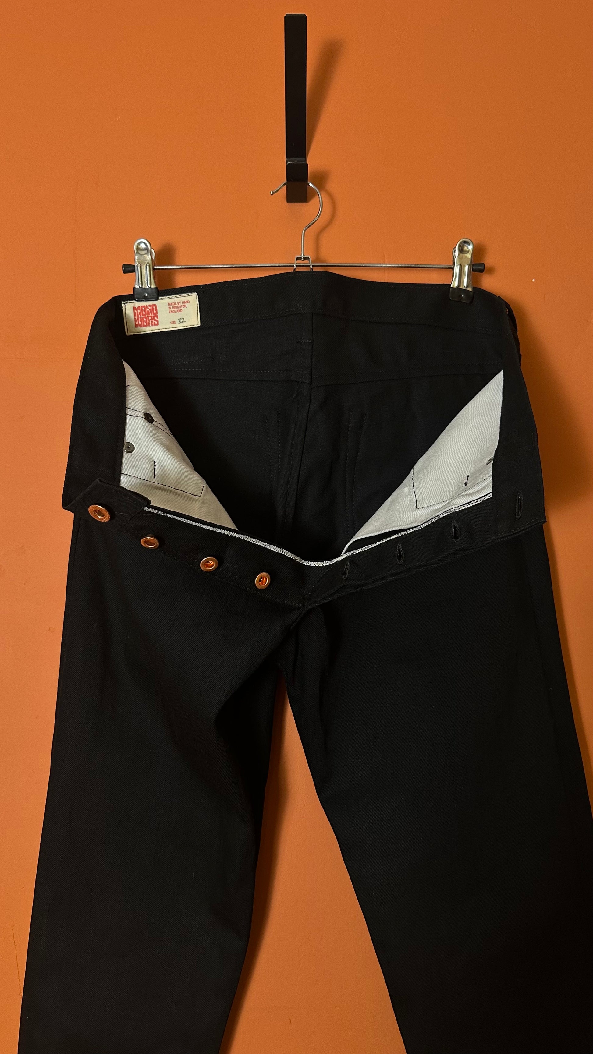 Black pants with brown buttons hanging on a rack against an orange wall