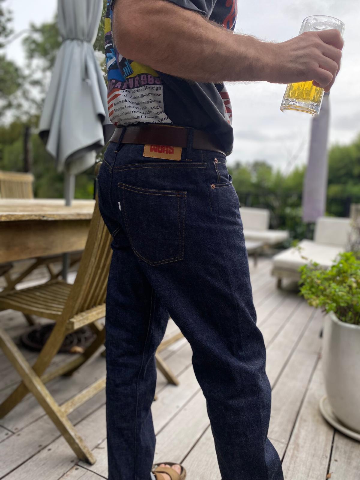 Person wearing dark blue jeans and a colorful shirt, holding a glass with a yellow label, on a wooden deck.