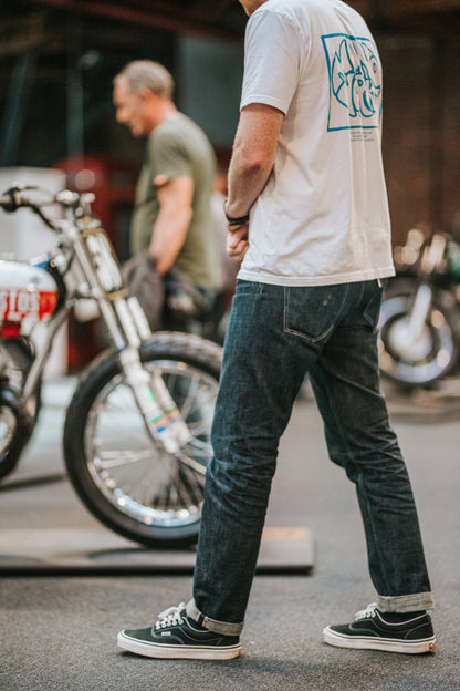 Person wearing a white t-shirt with a graphic design and dark jeans standing in front of motorcycles.